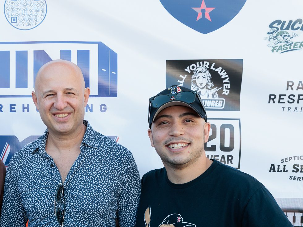Two guys smiling in front of the sponsor step and repeat at Fenway, with the Suck Faster logo in the top right corner