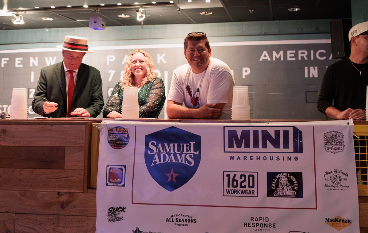 Ferg smiling at the judges' table at the Three Way Slider Championship, with the sponsor banner hanging in front and the Suck Faster logo in the bottom left